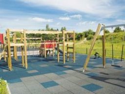 Holzspielgeräte mit Kletterturm, Schaukel und Rutsche, auf einem graublauem Spielplatzboden